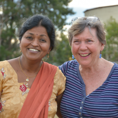 Creators of Peace South Asian woman in sari and white woman in blue tee-shirt stand shoulder to shoulder and smile at the camera