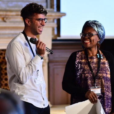 Initiatives for Land, Lives and Peace Young BIPOC man with glasses stands with microphone next to BIPOC woman with blue headwrap