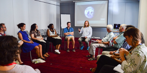 Group of mixed genders and ethnicities listen to a male with blonde hair speak in front of a presentation